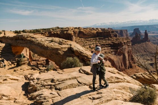 Two children hug on rocky terrain at a scenic overlook with dramatic sandstone arches and canyons in the background under a clear sky. by Justin Salem Meyer