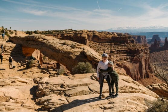 Two people stand on rocky terrain with a large natural stone arch behind them in a desert landscape. More people are visible in the background, and snow-capped mountains appear in the distance under a blue sky. by Justin Salem Meyer