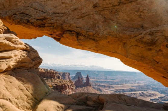 A natural red rock arch frames a panoramic desert landscape with mesas, canyons, and distant snow-capped mountains under a partly cloudy sky. by Justin Salem Meyer