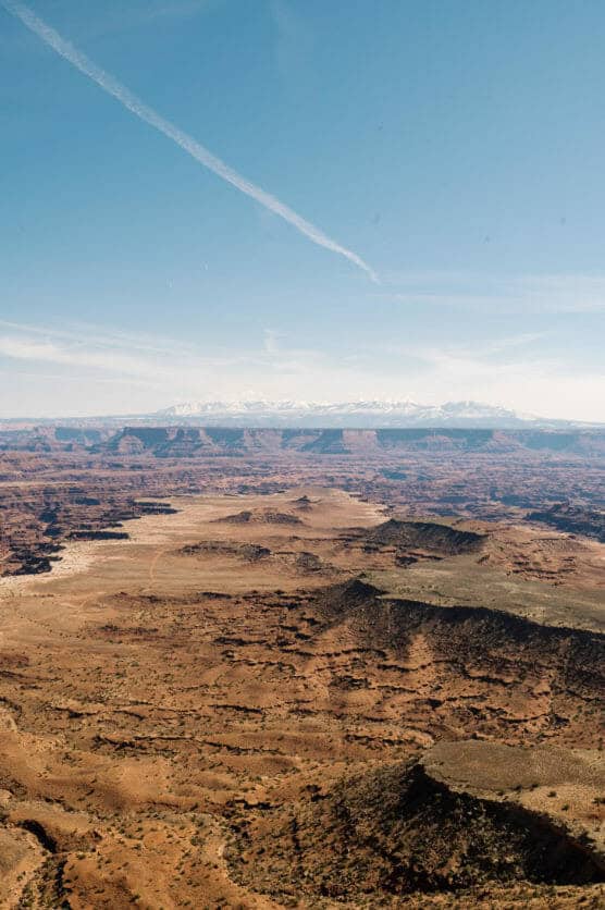 A vast, arid desert landscape with reddish-brown rocky terrain and mesas under a clear blue sky, with distant snow-capped mountains and a white contrail streaking across the sky. by Justin Salem Meyer