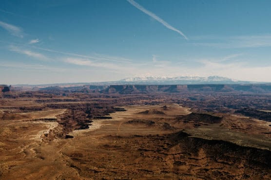 A vast, sunlit desert canyon with layered rock formations under a blue sky, distant snow-capped mountains on the horizon, and light clouds streaked across the sky. by Justin Salem Meyer