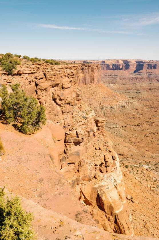 Steep rocky cliff edge overlooking a vast desert canyon landscape under a clear blue sky, with sparse green shrubs along the cliff. by Justin Salem Meyer
