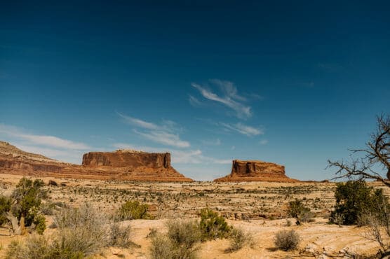 Desert landscape with two large, flat-topped red rock mesas under a deep blue sky, scattered clouds, and sparse desert vegetation in the foreground. by Justin Salem Meyer