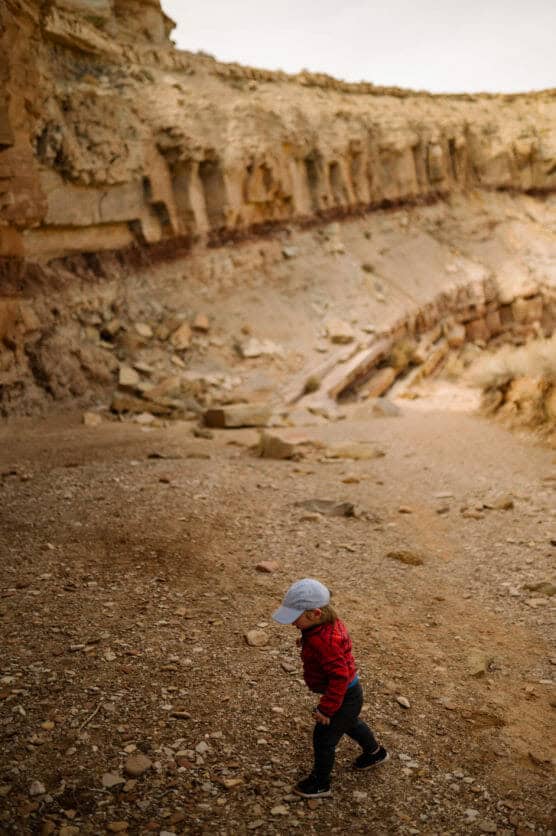 A young child wearing a red plaid shirt, blue pants, and a light blue cap walks alone on rocky ground in a canyon with steep, layered rock walls. by Justin Salem Meyer