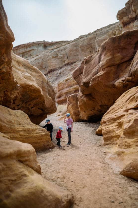 Three children walking on a sandy path between large, weathered rock formations in a desert canyon under a mostly cloudy sky. by Justin Salem Meyer