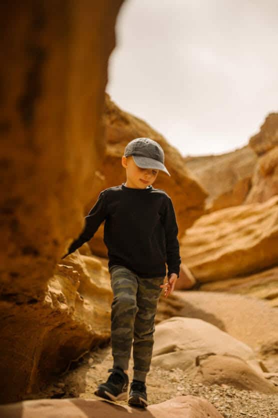 A young boy wearing a black cap, black sweatshirt, and camouflage pants walks along a rocky, sunlit canyon path, touching the canyon wall with one hand. The background features warm, orange rock formations. by Justin Salem Meyer