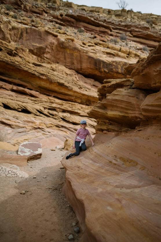 A child in a pink shirt and cap sits on layered, orange sandstone rocks in a desert canyon, with rugged, striated cliffs in the background. by Justin Salem Meyer