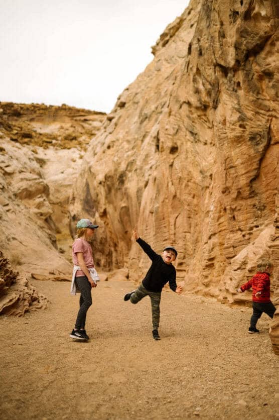 A woman and two children walk and play in a narrow canyon with towering tan rock walls. One child jumps with arms raised, while the other runs ahead. The setting appears dry and rugged. by Justin Salem Meyer