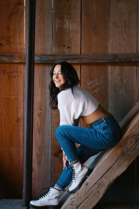 A woman with long dark hair, wearing a white cropped sweater, blue jeans, and white boots, sits sideways on wooden stairs, smiling over her shoulder in a rustic, wooden interior with handwritten notes on the wall. by Justin Salem Meyer