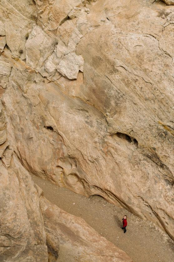 A person in a red jacket stands at the base of large, textured rock formations, emphasizing the vast scale and rugged surface of the surrounding landscape. by Justin Salem Meyer