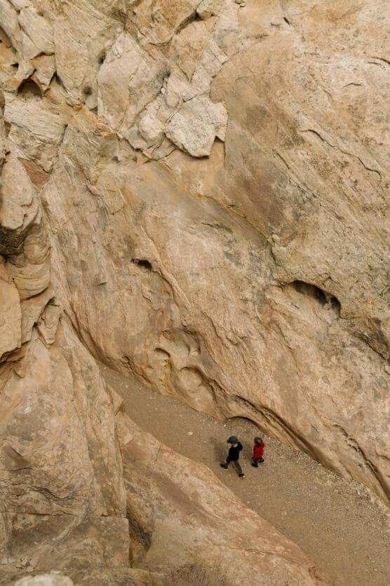 Two people walk along a narrow, sandy path at the base of towering, rugged rock walls in a canyon, viewed from above. The rocks are light brown and textured, creating a dramatic natural environment. by Justin Salem Meyer