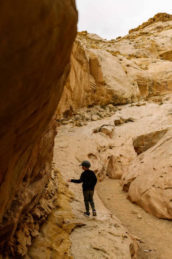 A young child wearing dark clothing walks along a narrow, rocky path surrounded by large, tan sandstone formations in a desert landscape. by Justin Salem Meyer