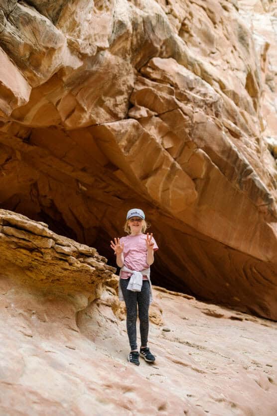 A child wearing a pink shirt, gray pants, and a cap stands on rocky ground in front of a large sandstone formation, holding up both hands with fingers spread wide. by Justin Salem Meyer