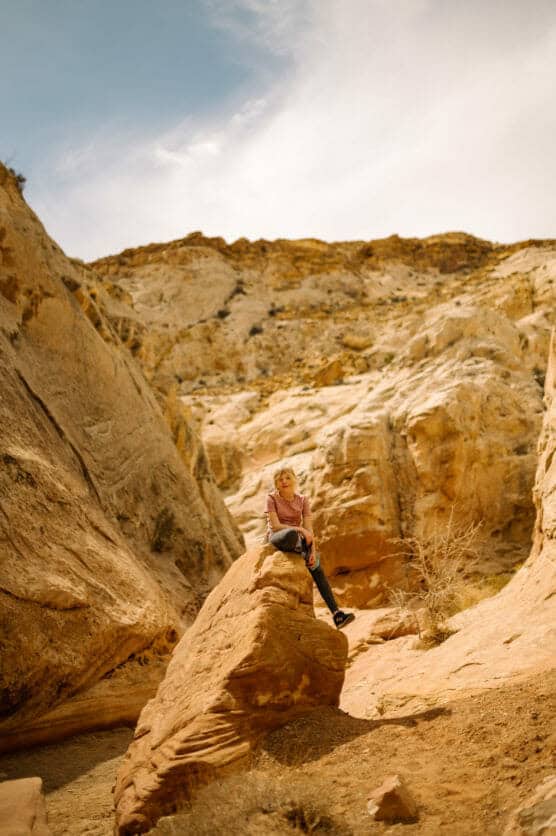 A person sits on a large red rock in a desert canyon, surrounded by rugged, sunlit sandstone cliffs under a partly cloudy sky. by Justin Salem Meyer