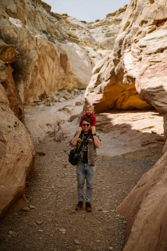 A man wearing sunglasses carries a smiling child on his shoulders while walking through a rocky desert canyon with beige and reddish stone walls. by Justin Salem Meyer