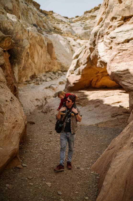 A man walks through a rocky canyon trail, carrying a child on his shoulders. The warm sunlight highlights the sandy path and textured rock formations surrounding them. by Justin Salem Meyer