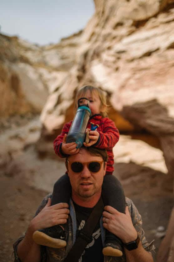 A man wearing sunglasses carries a young child on his shoulders through a rocky, sunlit canyon. The child, dressed in a red jacket, drinks from a blue water bottle. by Justin Salem Meyer