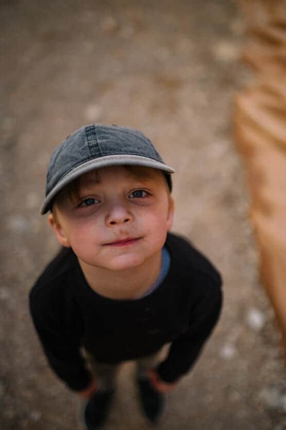 A young boy wearing a gray cap and black shirt looks up at the camera with a slight smile, standing outdoors on a dirt surface. The background is blurred. by Justin Salem Meyer