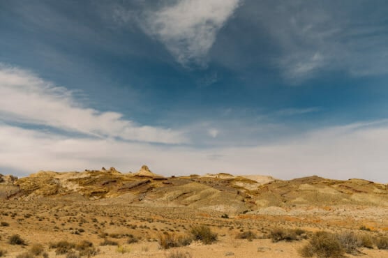 A wide desert landscape with rocky, tan-colored hills under a partly cloudy blue sky; sparse vegetation is scattered across the foreground. by Justin Salem Meyer