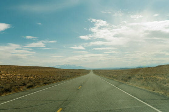 A long, empty road stretches straight into the distance through a dry, open landscape under a blue sky with scattered clouds and distant mountains on the horizon. by Justin Salem Meyer