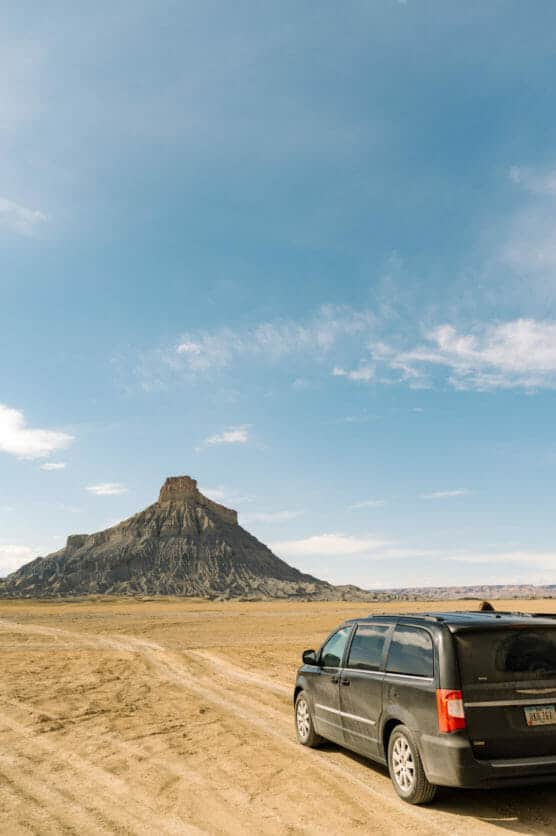 A black minivan parked on a dirt road in a desert landscape, with a flat-topped mesa rising in the background under a mostly clear, blue sky. by Justin Salem Meyer