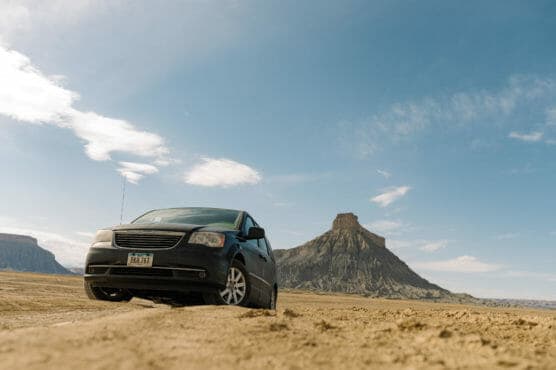 A black minivan parked on a sandy, desert landscape with a flat-topped mesa and blue sky in the background. by Justin Salem Meyer