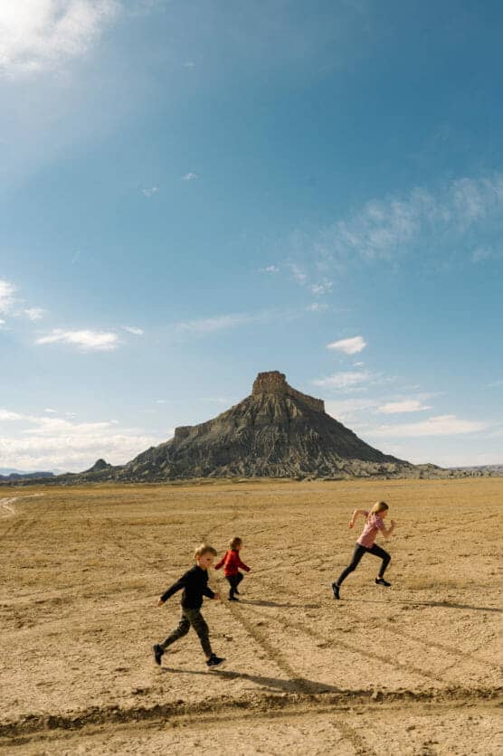 Three children run across a dry, sandy plain with a large, flat-topped mesa rising in the background under a bright blue sky with scattered clouds. by Justin Salem Meyer