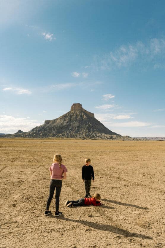 Three children stand and play on a dry, cracked desert landscape with a flat-topped mesa in the background under a clear blue sky. by Justin Salem Meyer