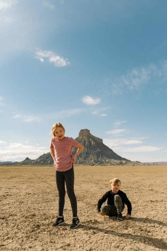 Two children are outdoors on a dry, cracked desert landscape with a flat-topped mountain in the background. The girl stands smiling, while the boy sits on the ground. The sky is bright and mostly clear. by Justin Salem Meyer