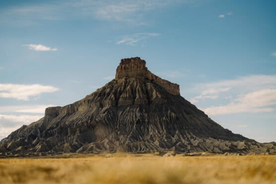 A flat-topped, rugged mesa rises from a barren, dry landscape under a blue sky with scattered clouds. The formation has steep, eroded sides with horizontal striations and sparse vegetation in the foreground. by Justin Salem Meyer
