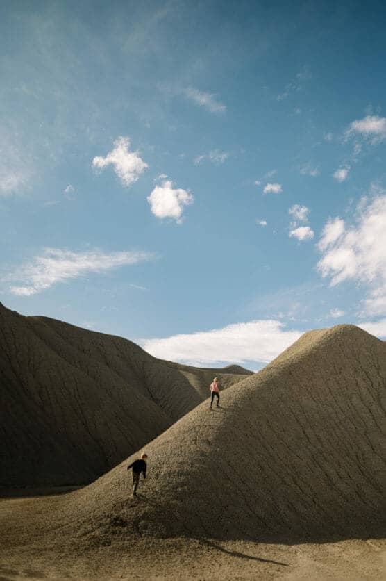 Two people climb separate barren, sandy hills under a blue sky with scattered clouds. The landscape appears dry and rugged, framed by larger hills in the background. by Justin Salem Meyer