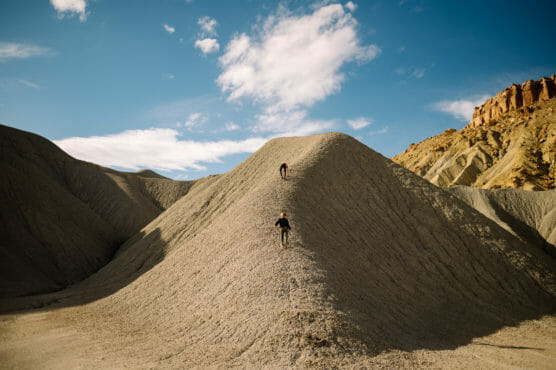 Two people climb up a steep, sandy hill surrounded by rugged, barren terrain under a blue sky with scattered clouds. Rocky cliffs are visible in the background. by Justin Salem Meyer