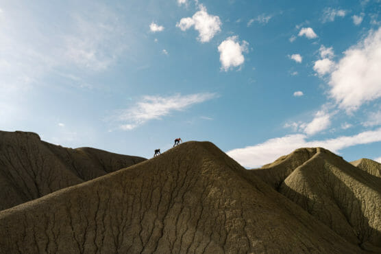 Three people climb a rugged, steep hill with cracked earth formations under a blue sky scattered with clouds. The figures appear small against the dramatic landscape. by Justin Salem Meyer