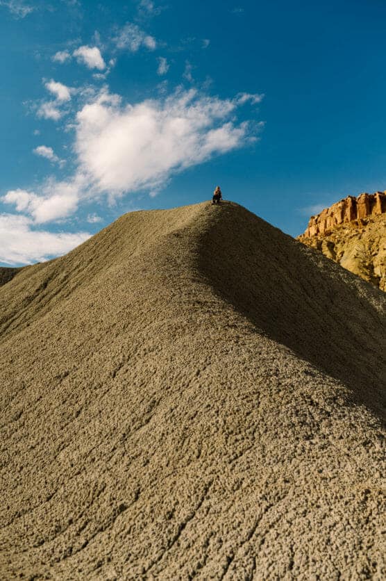 A person stands at the peak of a textured, rocky hill under a blue sky with scattered clouds, emphasizing the vast landscape and sense of solitude. by Justin Salem Meyer