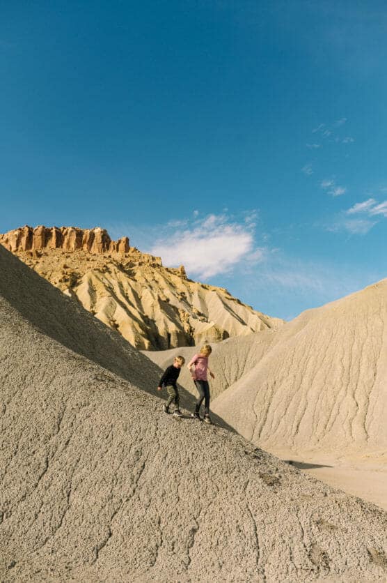Two people climb up a dry, rocky hill in a desert landscape with eroded beige formations and clear blue sky in the background. by Justin Salem Meyer
