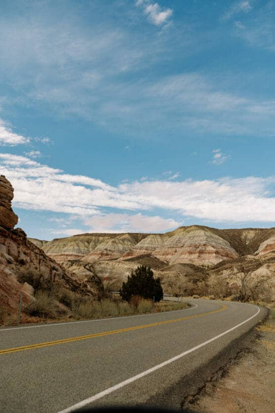 A winding road curves through a rocky desert landscape with layered hills and sparse vegetation under a partly cloudy blue sky. by Justin Salem Meyer