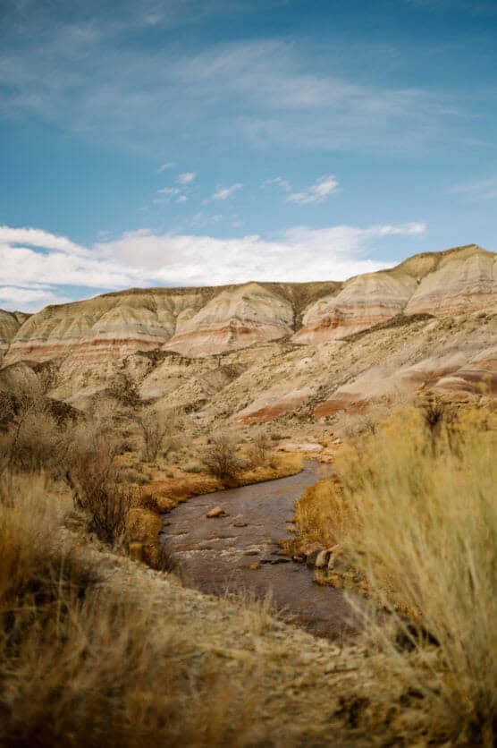 A small winding river flows through a dry, rocky landscape with layered hills in the background under a blue sky with scattered clouds. Sparse bushes and grasses line the riverbanks. by Justin Salem Meyer