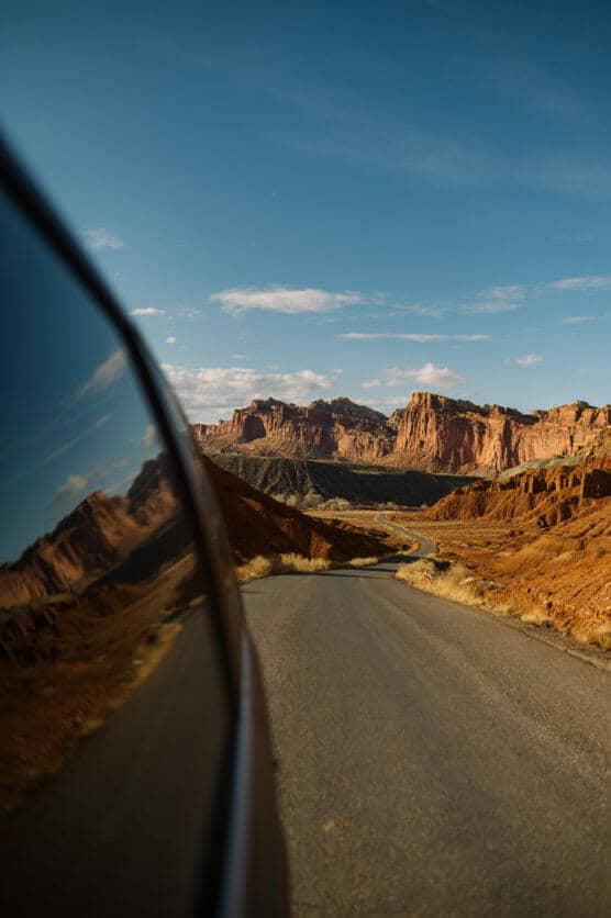 A paved road winds through a red rock desert landscape under a blue sky, with cliffs in the distance and a cars window reflecting the scenery on the left side of the image. by Justin Salem Meyer