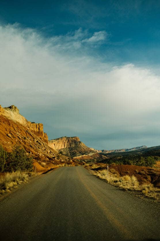 A paved road stretches through a desert landscape with red rock cliffs and sparse vegetation under a partly cloudy blue sky. by Justin Salem Meyer
