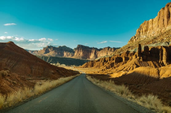 A paved road winds through a desert landscape with red rock formations and cliffs under a clear blue sky, with dry grass lining the roadside. by Justin Salem Meyer