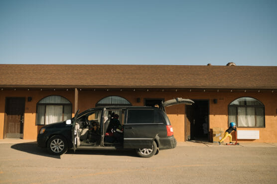 A black minivan with open doors is parked outside a tan motel. A person sits inside the van, and a child in a helmet rides a scooter nearby on the pavement under a clear blue sky. by Justin Salem Meyer