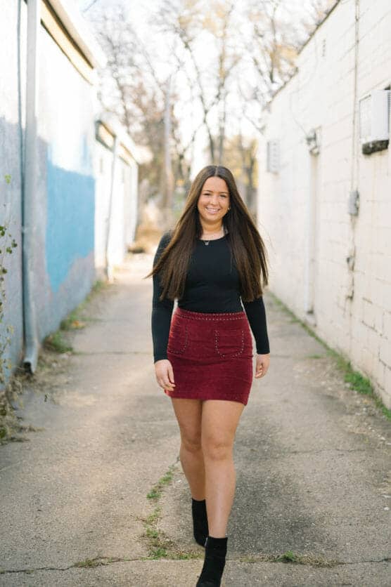 A young woman with long brown hair, wearing a black long-sleeve top, a red skirt, and black ankle boots, walks confidently down a sunlit alley with white and blue walls in the background. by Justin Salem Meyer