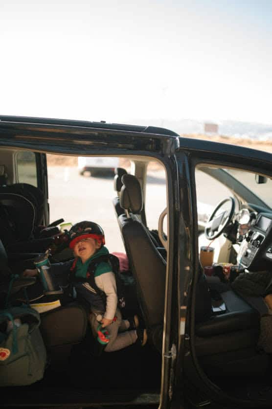 A toddler wearing a helmet sits in a car seat inside a parked vehicle with the sliding door open, holding a toy and a water bottle. Sunlight comes through the open door, and bags are visible next to the child. by Justin Salem Meyer