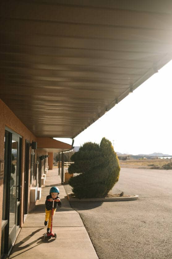 A child wearing a blue helmet rides a red scooter on a sunlit sidewalk next to a building, with bushes and an empty road in the background. by Justin Salem Meyer