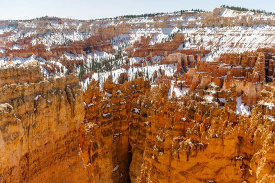 Orange and red rock formations with jagged spires rise from a canyon dusted with patches of snow, surrounded by forested areas under a clear sky. by Justin Salem Meyer