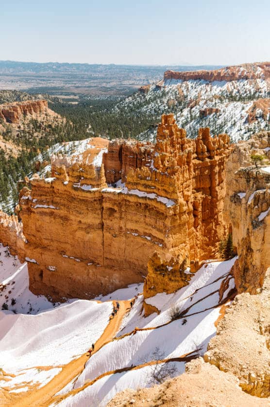 Tall orange rock formations rise above snowy slopes and pine trees in a canyon landscape under a clear sky, likely in Bryce Canyon National Park. by Justin Salem Meyer