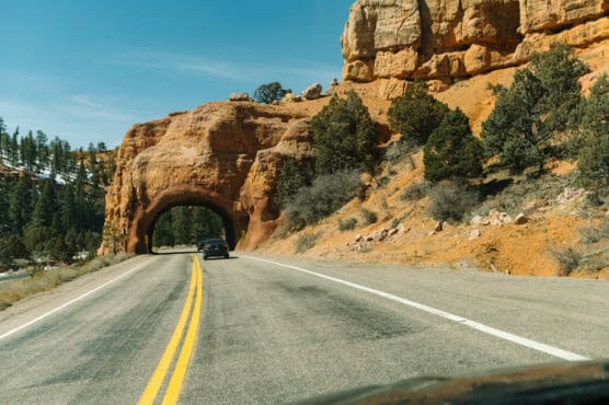 A car drives through a natural red rock tunnel on a scenic road surrounded by rocky cliffs and green trees under a clear blue sky. by Justin Salem Meyer