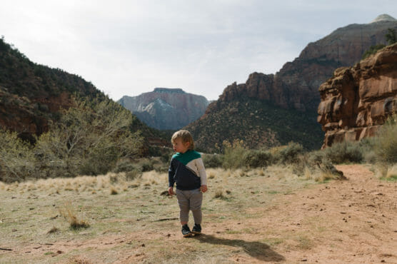 A young child stands on a dirt trail surrounded by rugged mountains and sparse vegetation under a partly cloudy sky in a scenic outdoor landscape. by Justin Salem Meyer