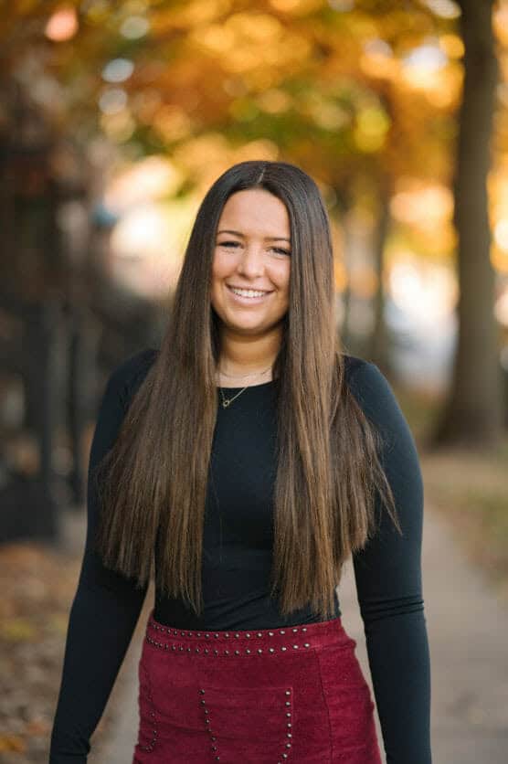 A woman with long straight brown hair, wearing a black top and a maroon skirt, stands outdoors smiling with autumn trees and fallen leaves in the background. by Justin Salem Meyer