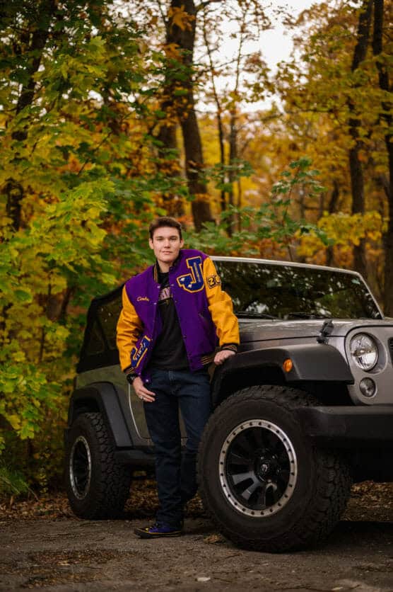 A young man in a purple and yellow varsity jacket stands beside a gray Jeep in a wooded area with autumn foliage. He is smiling and posing with one hand in his pocket. by Justin Salem Meyer